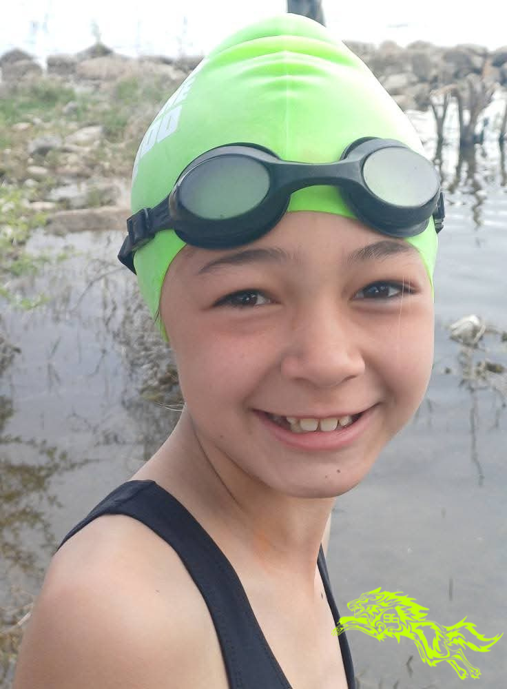 Ten-year-old Grace smiling at the camera wearing a bright green swimming cap and goggles, standing at the water's edge