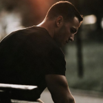 Muscular man sitting alone on a park bench at dusk, head down, amber light behind him — representing chronic stress and elevated cortisol