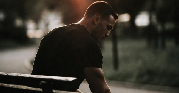 Muscular man sitting alone on a park bench at dusk, head down, amber light behind him — representing chronic stress and elevated cortisol